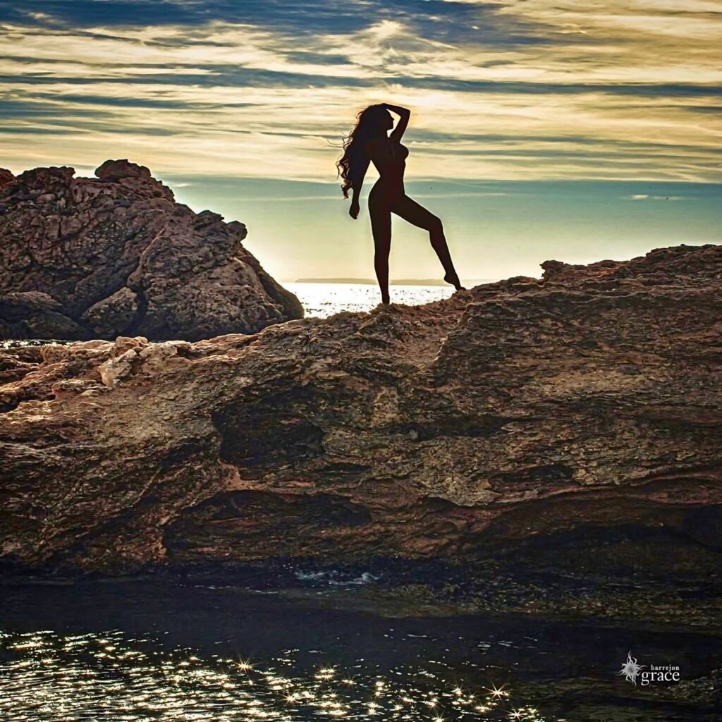 Silueta de una mujer desnuda en una roca al atardecer, con el mar y el cielo en el fondo