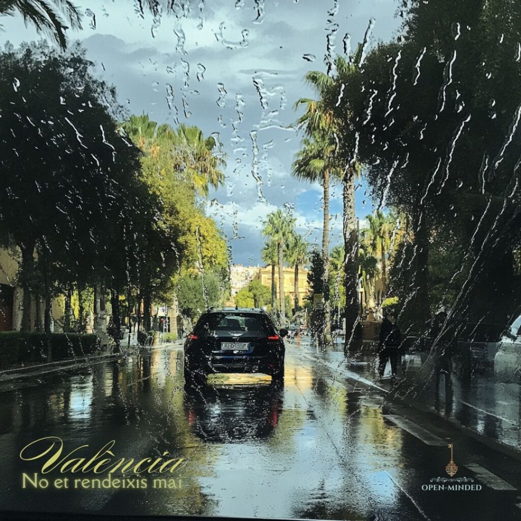 Vista de una calle en Valencia bajo la lluvia, con gotas de agua en el parabrisas que distorsionan la imagen de palmeras y un cielo nublado. Texto en la esquina inferior izquierda: ‘València, No et rendeixis mai.’ Logo de OPEN-MINDED en la esquina inferior derecha